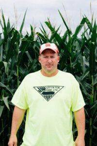 Man wearing an orange and white hate and a neon yellow JWV shirt in front of a corn field