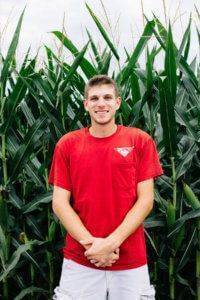 Young adult man standing in front of corn field holding his hands while wearing a bright red JWV t-shirt